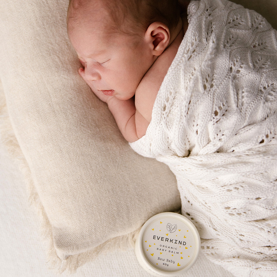 Photo of Everkind's New Baby balm next to a sleeping baby. A night time massage with New Baby is the perfect way to settle a newborn, especially in those first few months.